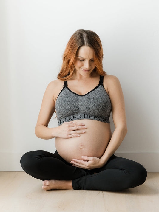 Pregnant model sitting on the ground holding her pregnant belly while wearing the Sublime® Nursing Sports Bra in Heather Grey @model_info:Shannon is wearing a Small Busty.