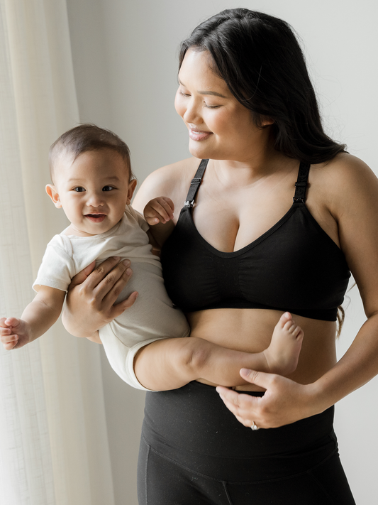 Model holding her baby while wearing the Sublime® Hands-Free Pumping & Nursing Sports Bra in Black @model_info:Binc is wearing a Medium Busty.